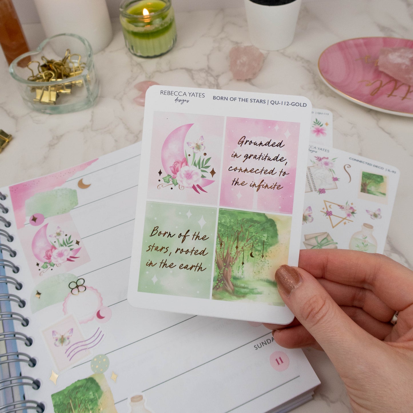 Hand holding a gold-accented sticker sheet with celestial moon and woodland designs, surrounded by a lit candle and journaling supplies.