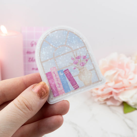 Hand holding a decorative sticker with a window design featuring books and flowers on a soft background.
