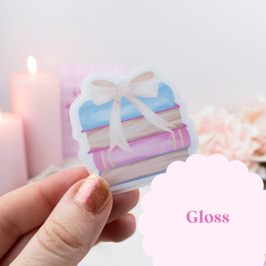 Hand holding a sticker of books with a bow, with 'Gloss' text in the corner.