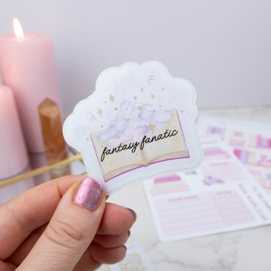 Hand holding a decorative sticker with 'fantasy fanatic' text, pink candle in the background