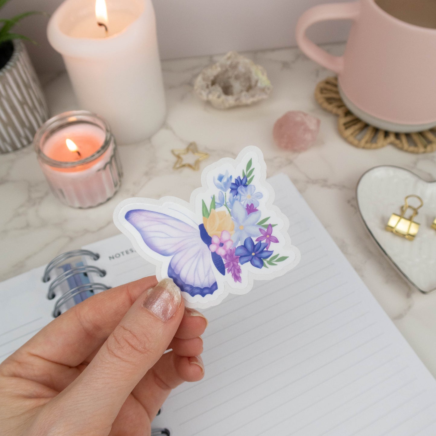 Hand holding a floral butterfly sticker with candles and a notebook in the background