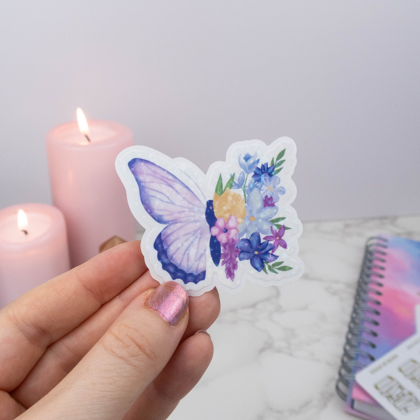 Butterfly-shaped sticker with floral design held by a hand, with candles and a notebook in the background.