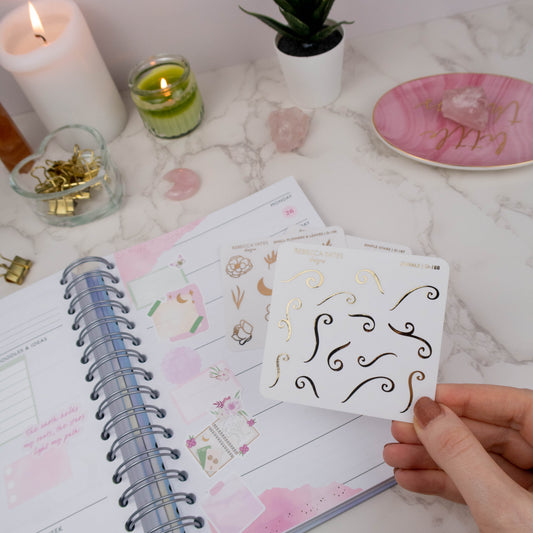 Hand holding a transparent matte sticker sheet with decorative swirl designs in gold foil, surrounded by journaling supplies and a planner.