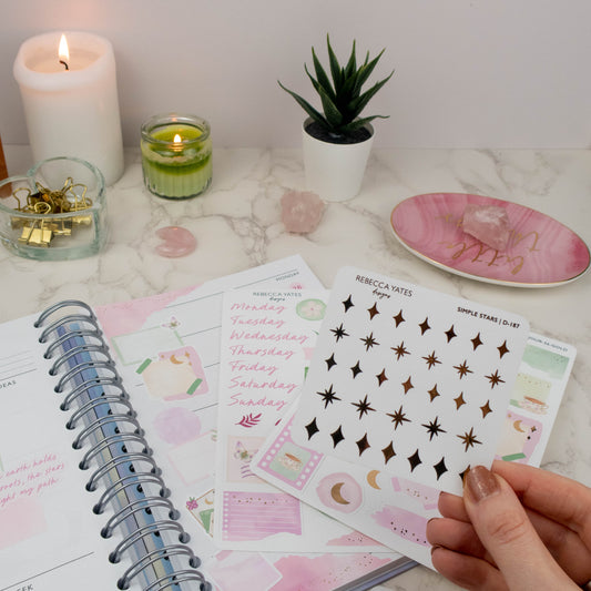 Hand holding a celestial sticker sheet with simple star designs in gold foil, surrounded by journaling supplies and a planner."