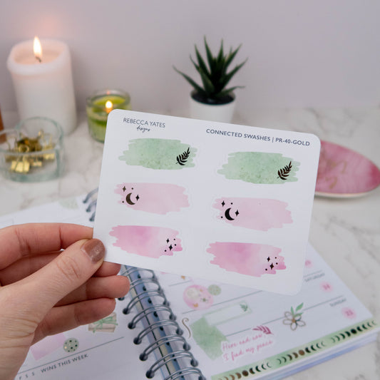 Hand holding a transparent matte sticker sheet with gold-foiled swash box designs in pink and green tones, surrounded by journaling supplies.