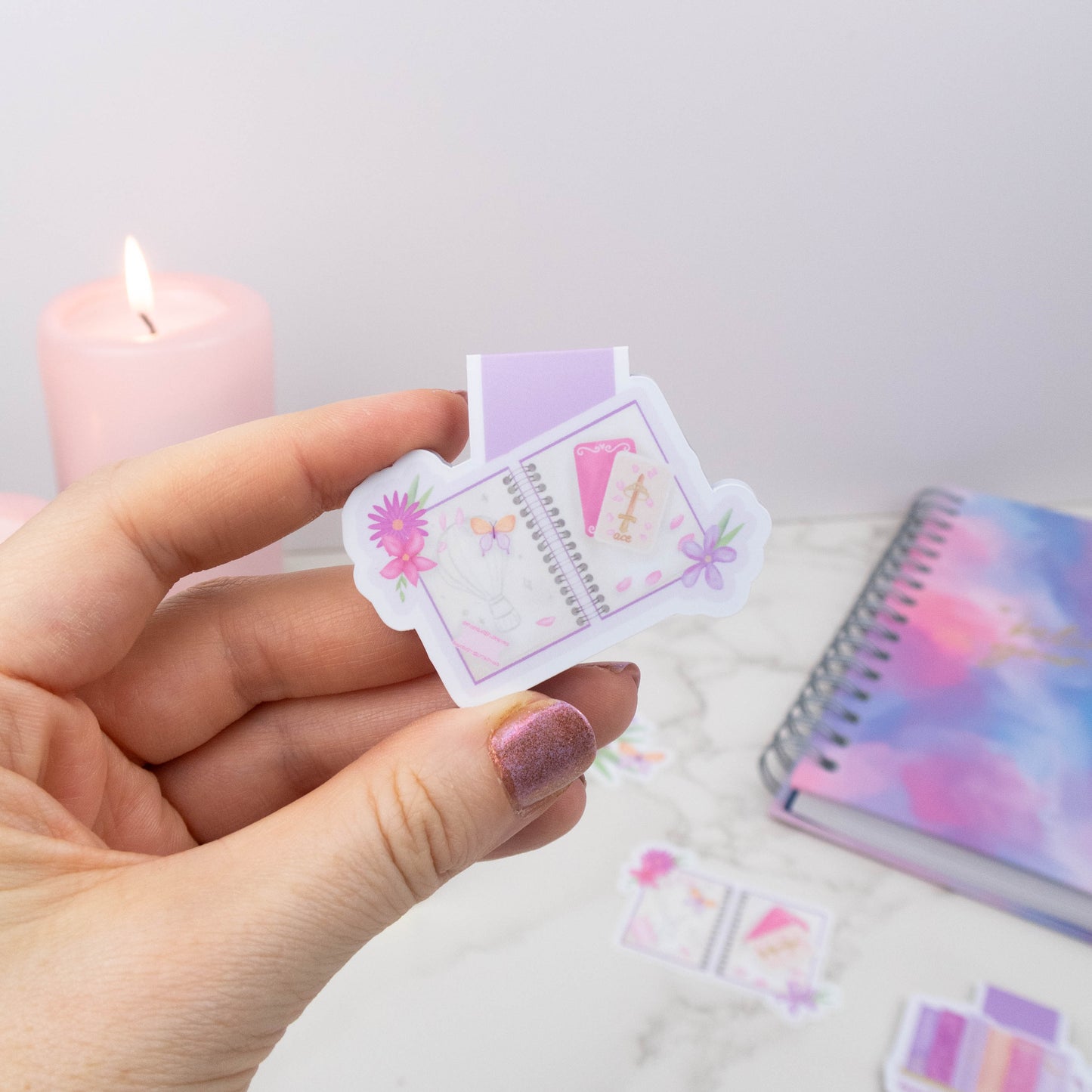 Hand holding a small, colorful notebook with a candle and notebook in the background