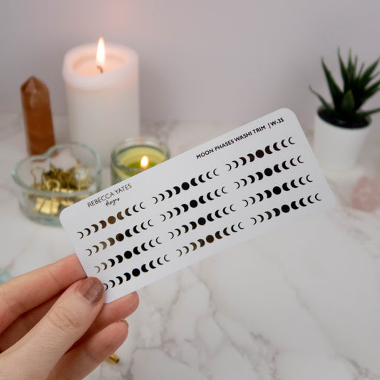 Moon Phases Washi Style Trim Sticker Sheet featuring foiled lunar phase designs in gold, displayed on a marble desk with a lit candle and stationery