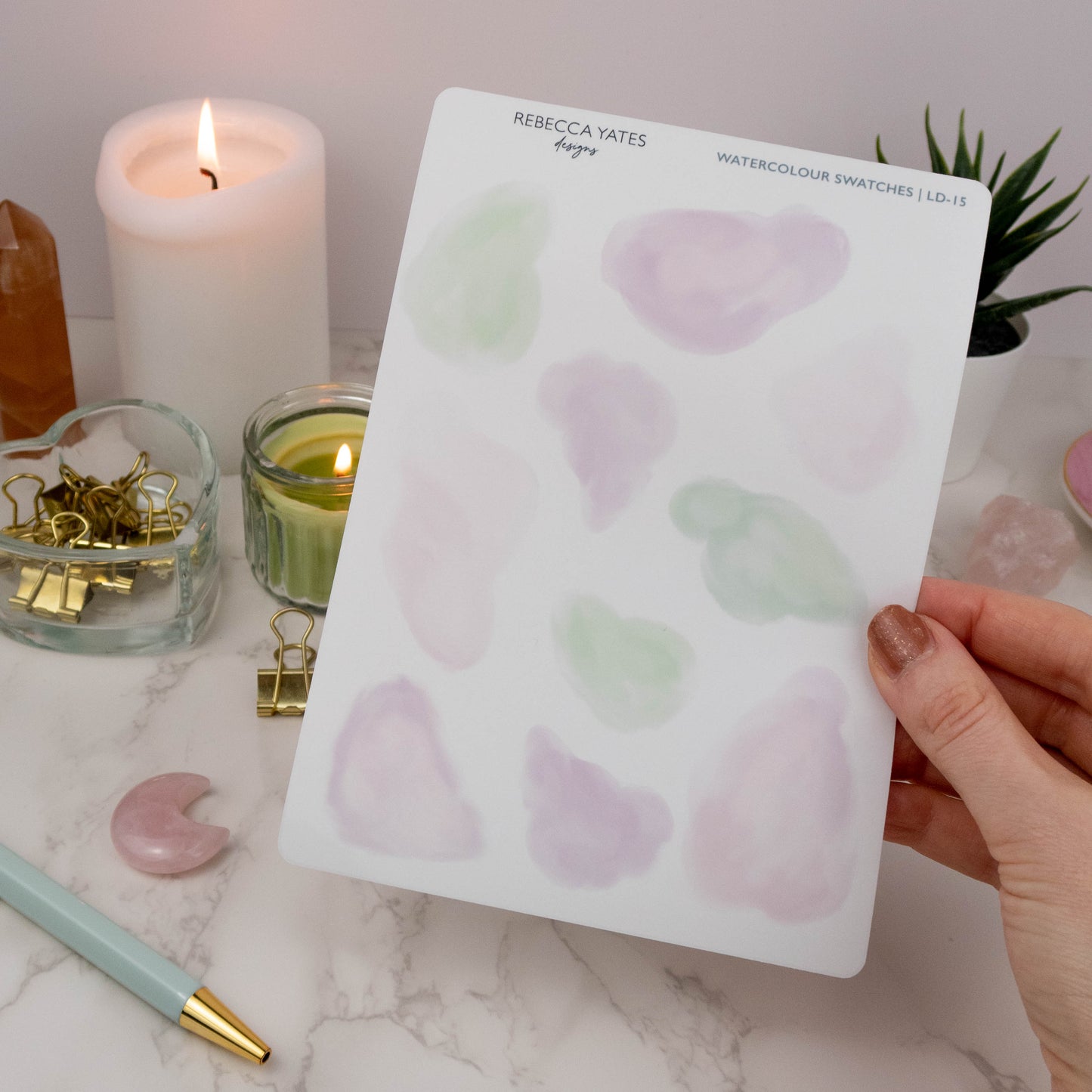 Hand holding a watercolour swatch sticker sheet with pink and green tones, ideal for planners and bullet journals, surrounded by journaling supplies