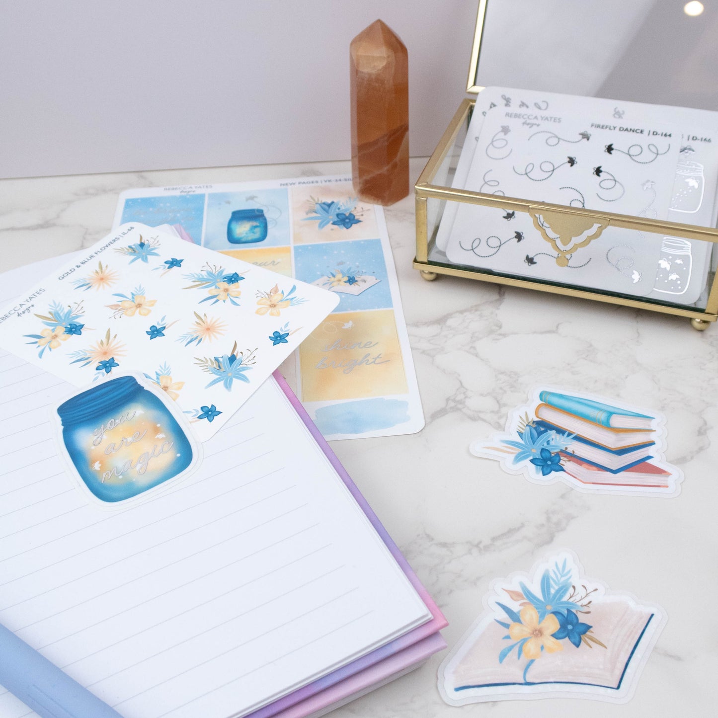 Sticker sheets with decorative designs on a marble surface next to a jewelry box.