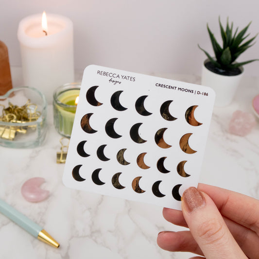 Crescent Moon Sticker Sheet featuring 28 foiled designs in gold, displayed on a marble desk with a lit candle and stationery.
