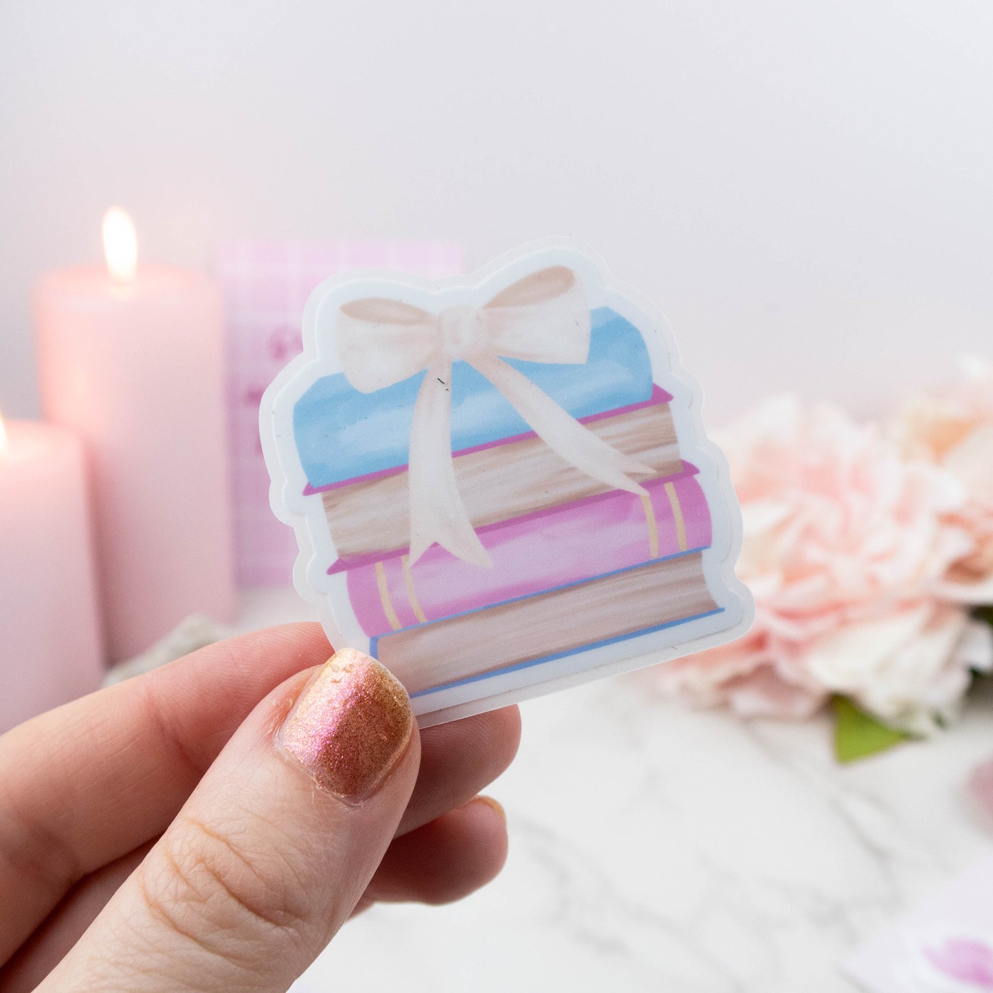 Hand holding a decorative sticker of books with a bow, with candles and flowers in the background.