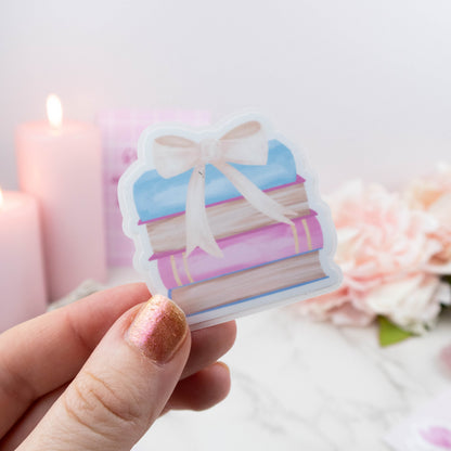 Hand holding a decorative sticker of books with a bow, with candles and flowers in the background.