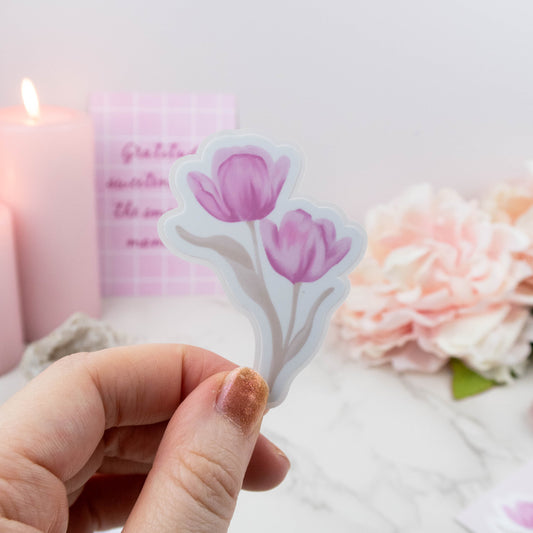Hand holding a tulip-shaped sticker with a blurred candle and flowers in the background