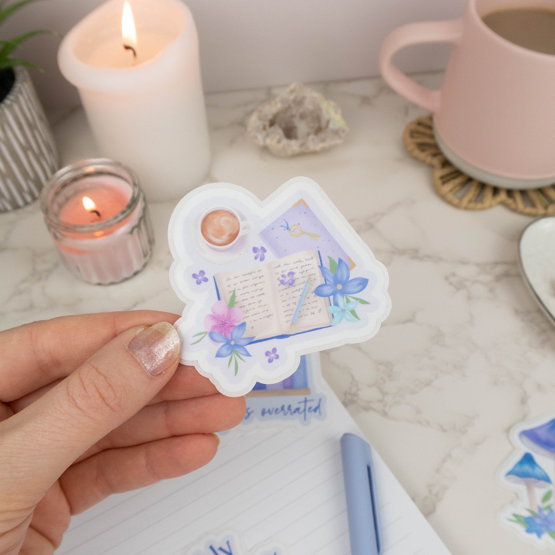 Hand holding a decorative sticker with a book and flower design on a table with candles and a mug.