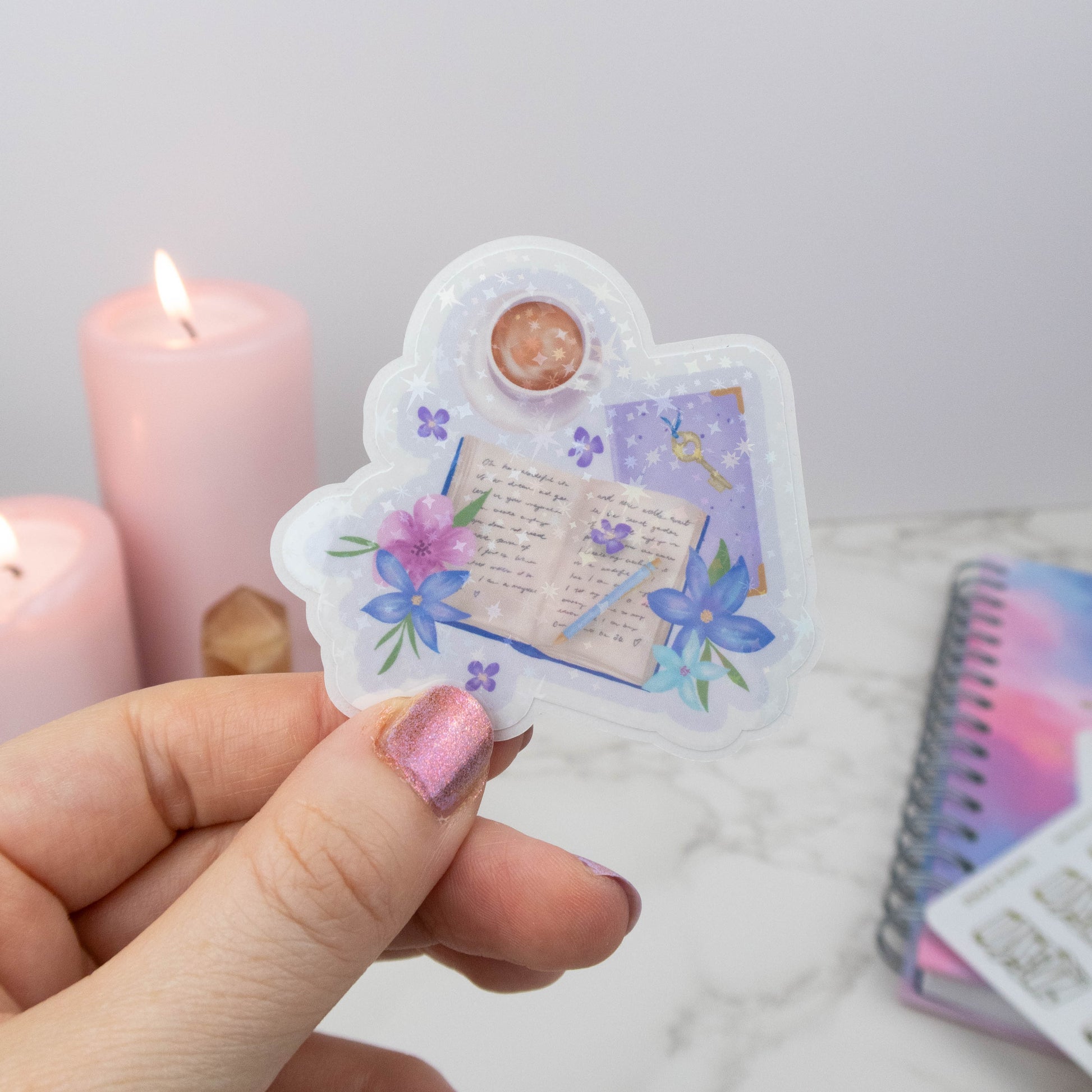 Hand holding a decorative sticker with floral and text design, candles and notebook in the background.