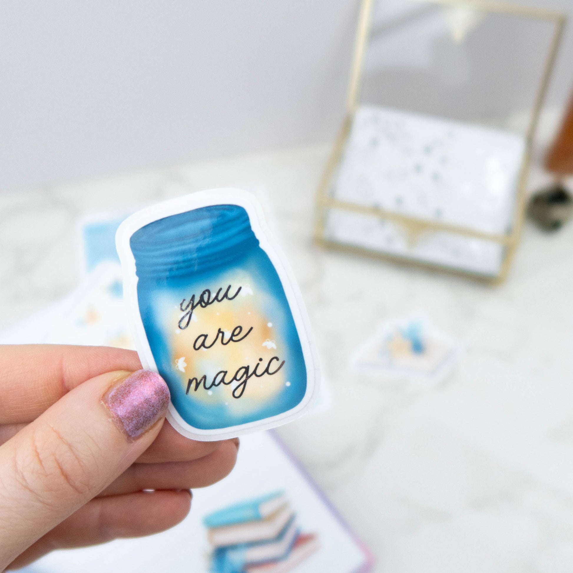 Hand holding a blue jar sticker with 'you are magic' text. jar is filled with fireflies 
