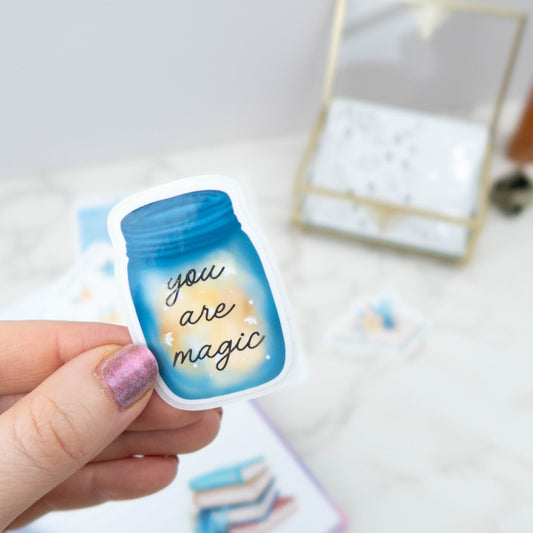 Hand holding a blue jar sticker with 'you are magic' text. jar is filled with fireflies 