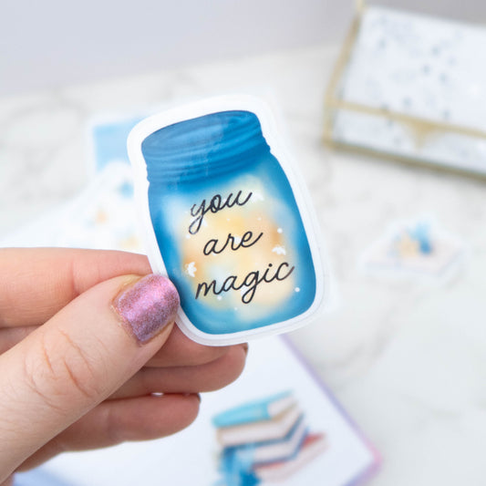 Hand holding a small blue jar with 'you are magic' text on a blurred background