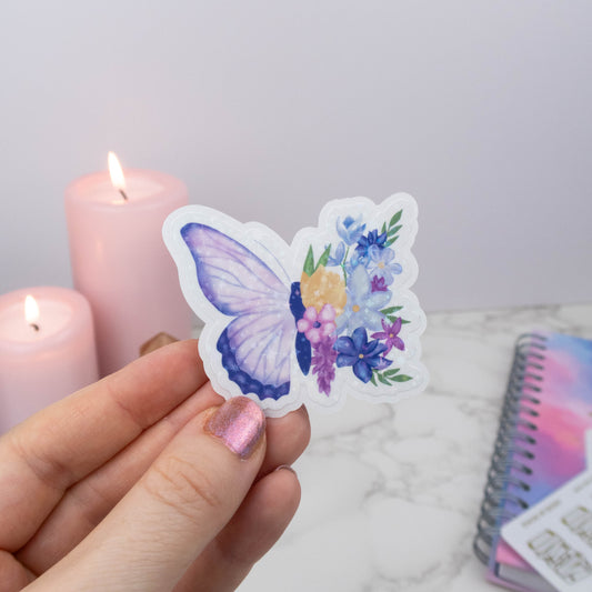Butterfly-shaped sticker with floral design held by a hand, with candles and a notebook in the background.