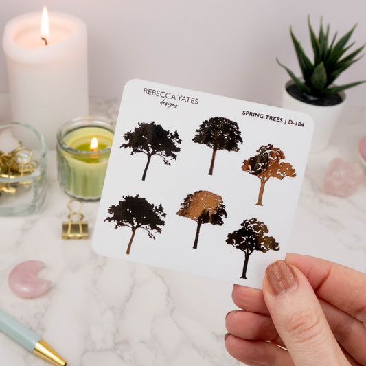 Hand holding a transparent matte sticker sheet with spring tree designs in gold foil, surrounded by journaling supplies and a planner.