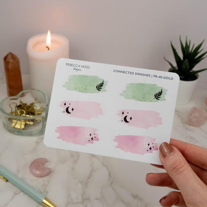 Swash Box Sticker Sheet featuring six pink and green watercolour swash designs with gold foil details, displayed on a marble desk with a lit candle and planner.