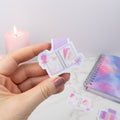 Hand holding a small, colorful notebook with a candle and notebook in the background