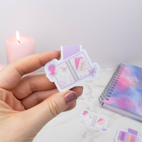 Hand holding a small, colorful notebook with a candle and notebook in the background