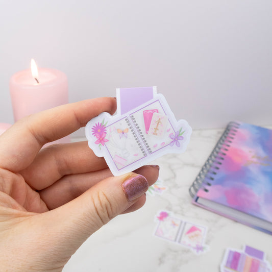 Hand holding a small, colorful notebook with a candle and notebook in the background