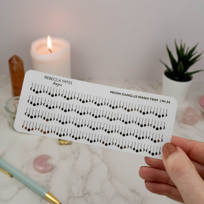 Moon Dangles Washi Style Trim Sticker Sheet featuring foiled moon designs in gold, displayed on a marble desk with a lit candle and stationery.