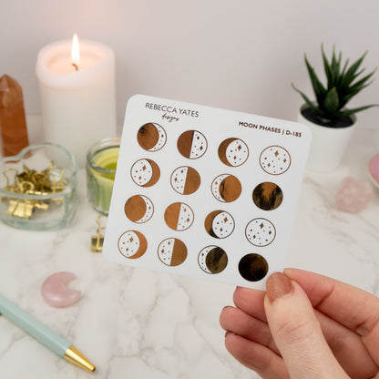 Moon Phases Sticker Sheet featuring 18 foiled designs for tracking the lunar cycle, displayed on a marble desk with a lit candle and stationery.