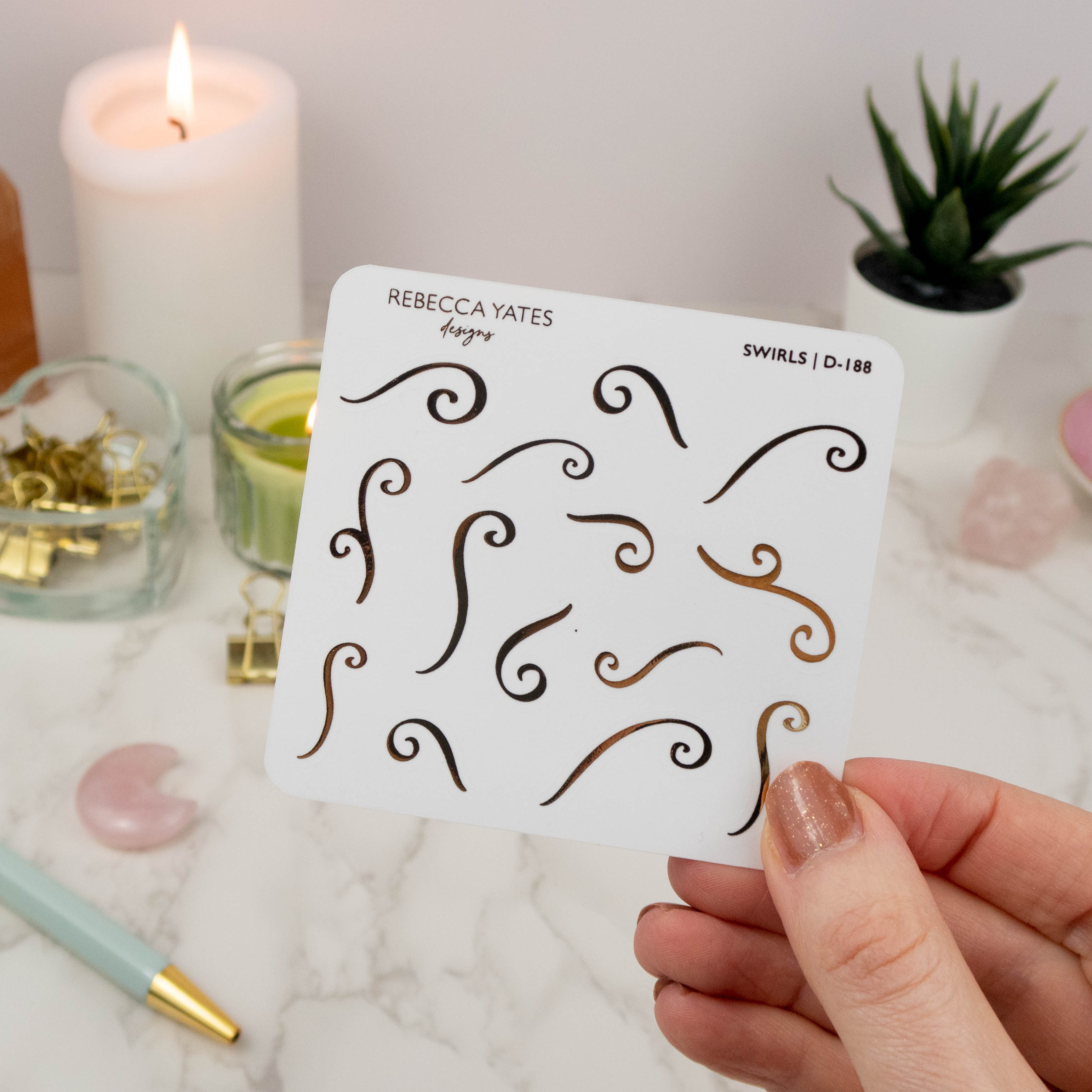 Swirls Sticker Sheet featuring 24 foiled designs in gold, displayed on a marble desk with a lit candle and stationery."