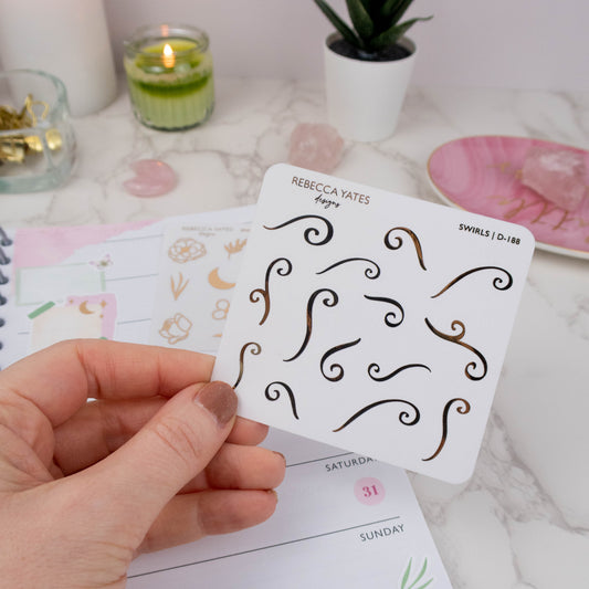 Swirls Sticker Sheet featuring 24 foiled designs in gold, displayed on a marble desk with a lit candle and stationery.