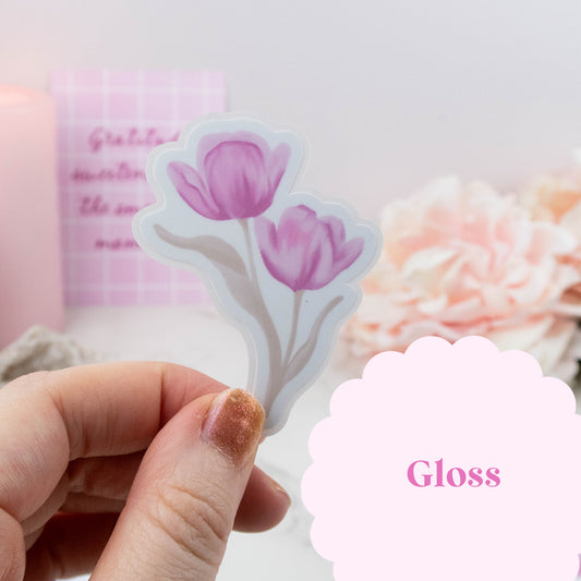 Hand holding a floral sticker with 'Gloss' text, blurred flowers in the background