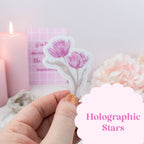 Hand holding a floral sticker with pink flowers, blurred background with candles and flowers.